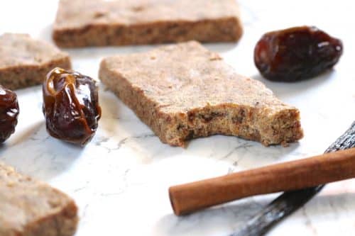 A thin protein bar with a bite missing in the focus of the shot surrounded by more bars, whole dates, a cinnamon stick, and a vanilla bean.