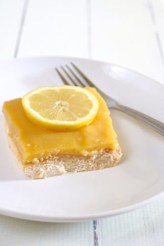A lemon bar on a white plate with a fork in the background.