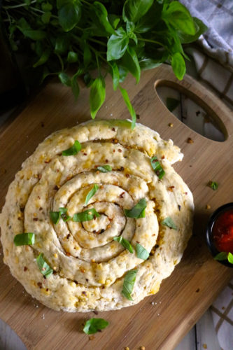 A crust rolled into a spiral topped with torn basil and red pepper flakes. A large bunch of basil is towards the top of the photo.