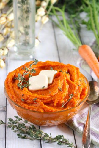 A glass bowl of mashed carrots topped with two pats of butter and fresh thyme. Carrots, flowers, and more thyme in a glass bottle are in the background.