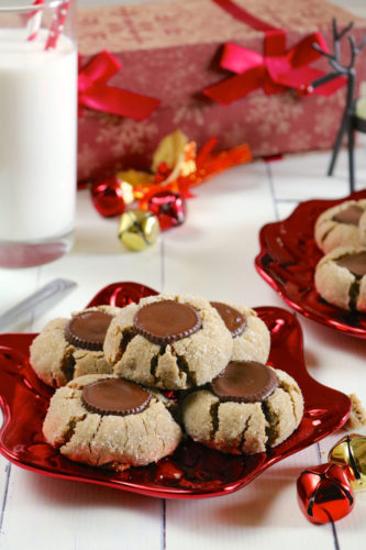 A red plate of sunbutter blossom cookies with mini peanut butter cups in the center of each and Christmas decorations in the background.