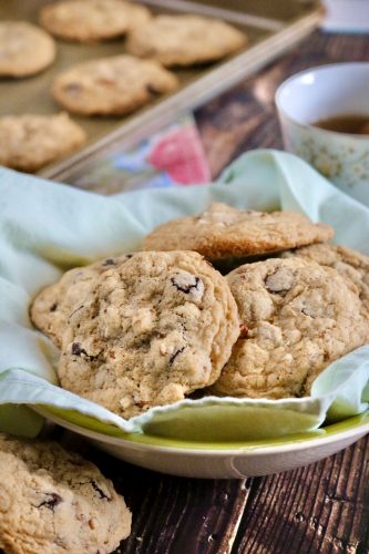 Chocolate chip cookies in a linen lined bowl. An out of focus background shows a cup of tea and additional cookies on a baking sheet all on a wood background.