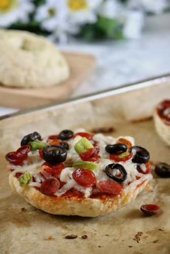 A single bagel pizza still on the parchment lined baking sheet with a bagel half and flowers faded in the background.