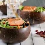 Two bowls of salmon salad, one in focus, and a spoonful of dried cranberries off to the right. Flowers and sparkling water are barely visible in the background.