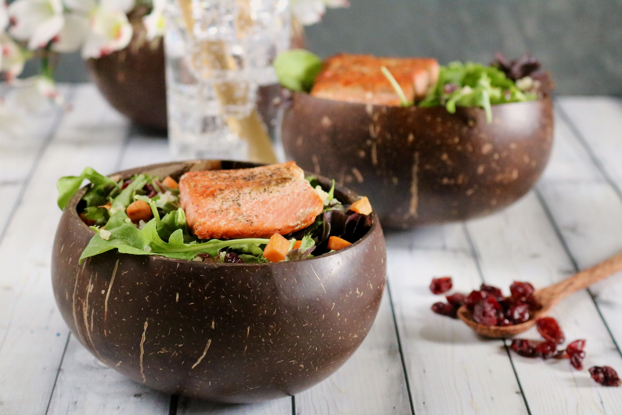 Two bowls of salmon salad, one in focus, and a spoonful of dried cranberries off to the right. Flowers and sparkling water are barely visible in the background.