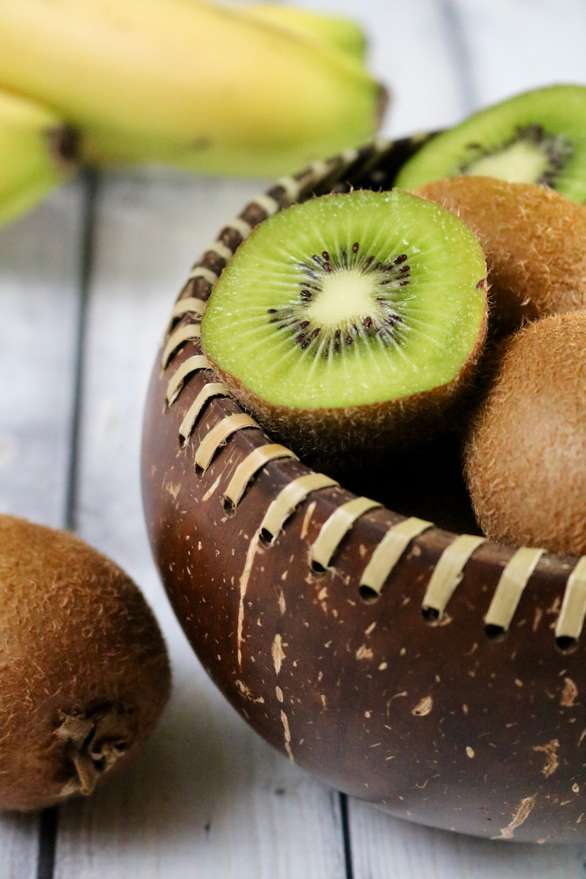 Fiber laced coconut shell bowl being used to hold kiwi with bananas in the background.