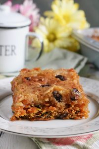 Up close side view of a square slice of gooey butter cake on a white plate. Flowers and a white cup with lid are in the background.
