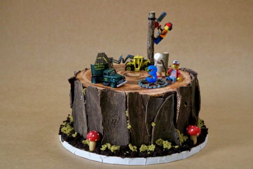 A cake decorated to look like a tree stump with moss, dirt, and mushrooms around the outside and topped with a logging scene made using Lego people and toy trucks.