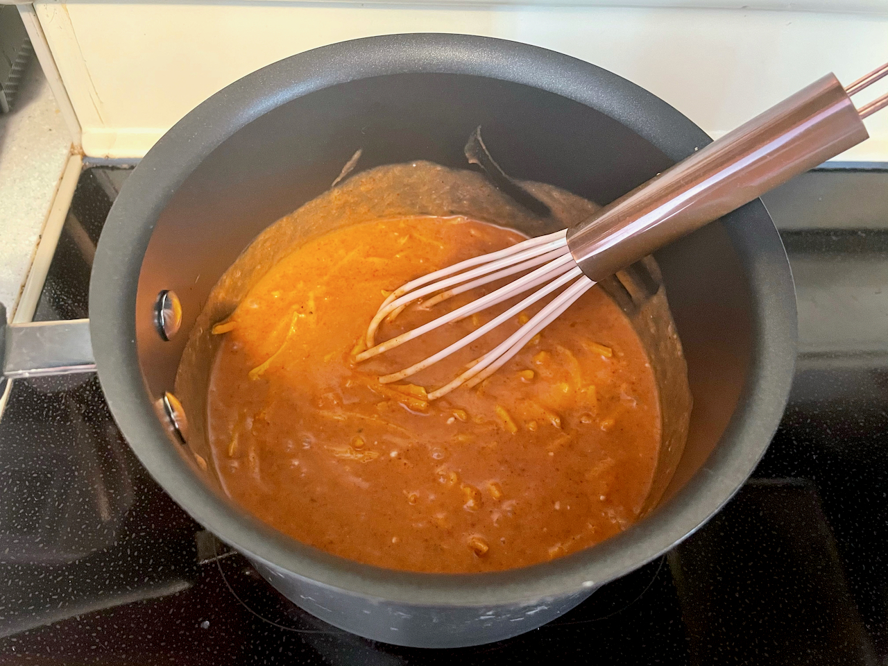 Orange sauce with visible cheese shreds in a black pot with a whisk in it.