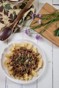 A plate of stroganoff over noodles next to a wooden spoon and chives with flowers still attached.